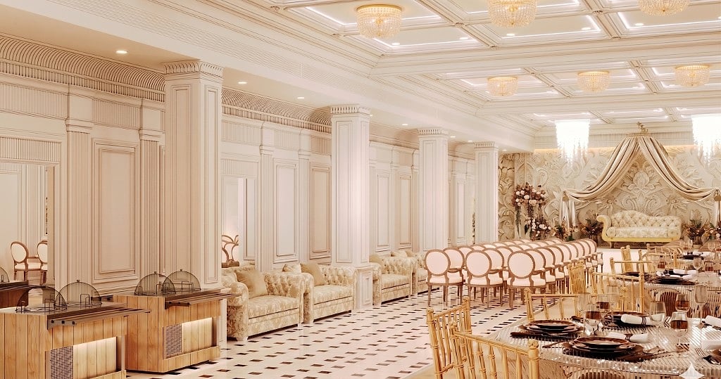 The banquet hall along its length, tufted cream lounge sofas and fluted columns, warm-wood buffet counters, gold-chiavari banquet tables and a draped stage at the far end