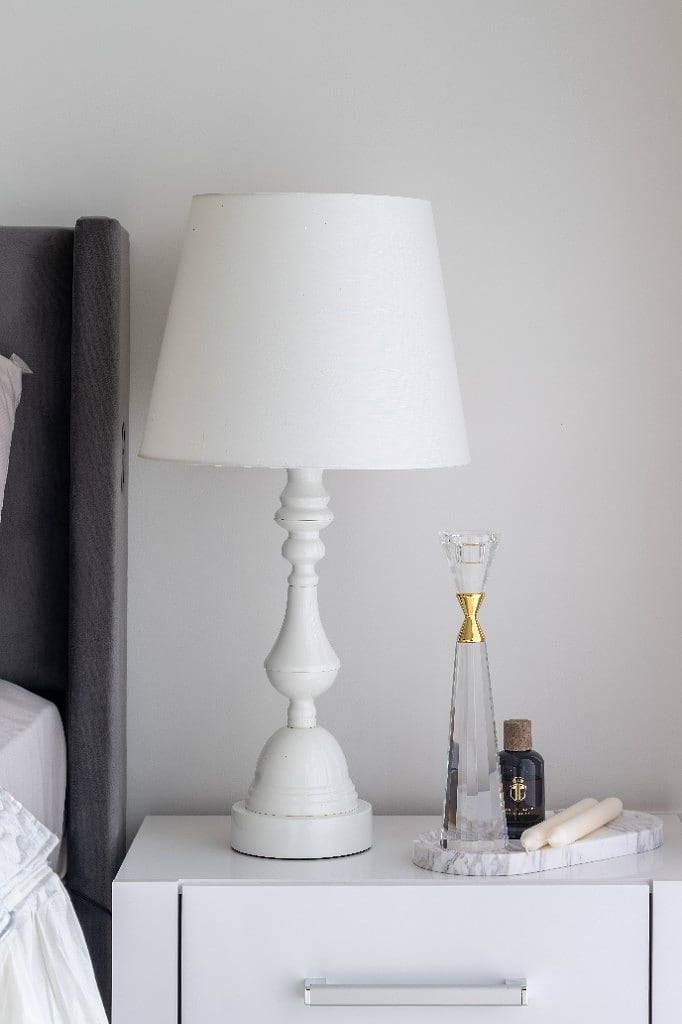 Bedside detail, a white turned-wood lamp beside a marble tray holding a crystal candlestick with a brass collar, set against the grey channel-tufted headboard