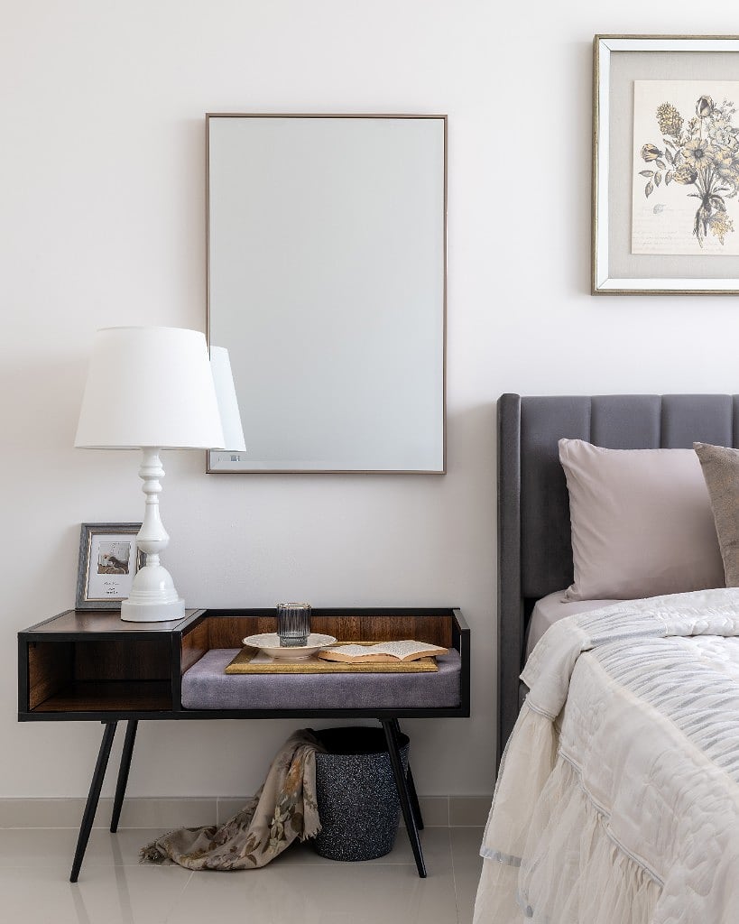Dressing corner with a bevelled mirror above a walnut-and-black console, a white lamp and a framed photograph, beside a silver-framed botanical print and the grey tufted bed