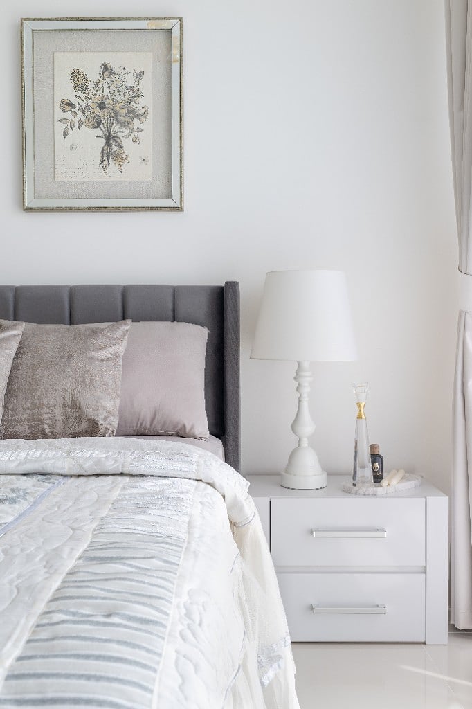 Bedside vignette, a single silver-framed botanical print above a white nightstand styled with a turned-wood lamp, a crystal candlestick and a marble tray, against the grey velvet headboard
