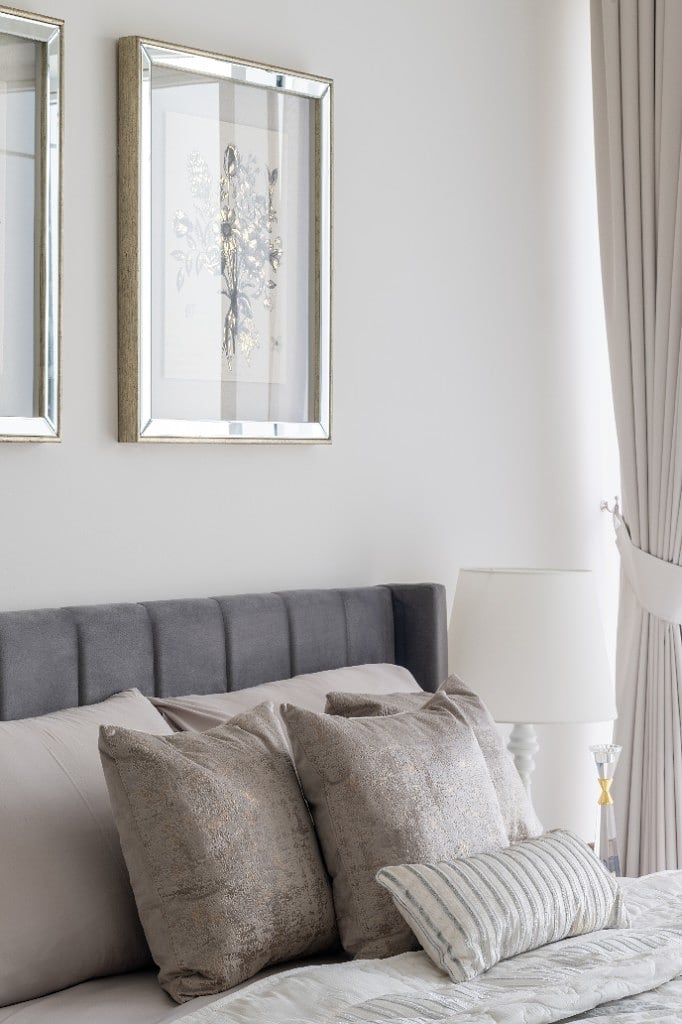 A pair of mirror-framed botanical prints above the grey channel-tufted headboard, dressed with taupe velvet cushions and a soft pleated curtain held back with a tieback
