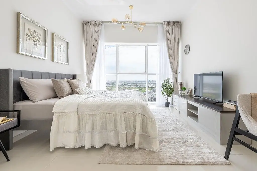 Floor-to-ceiling window onto the Damac Hills golf course behind a grey upholstered bed, with sheer linen and taupe drapery, a gold glass chandelier and a white media console carrying a trailing plant
