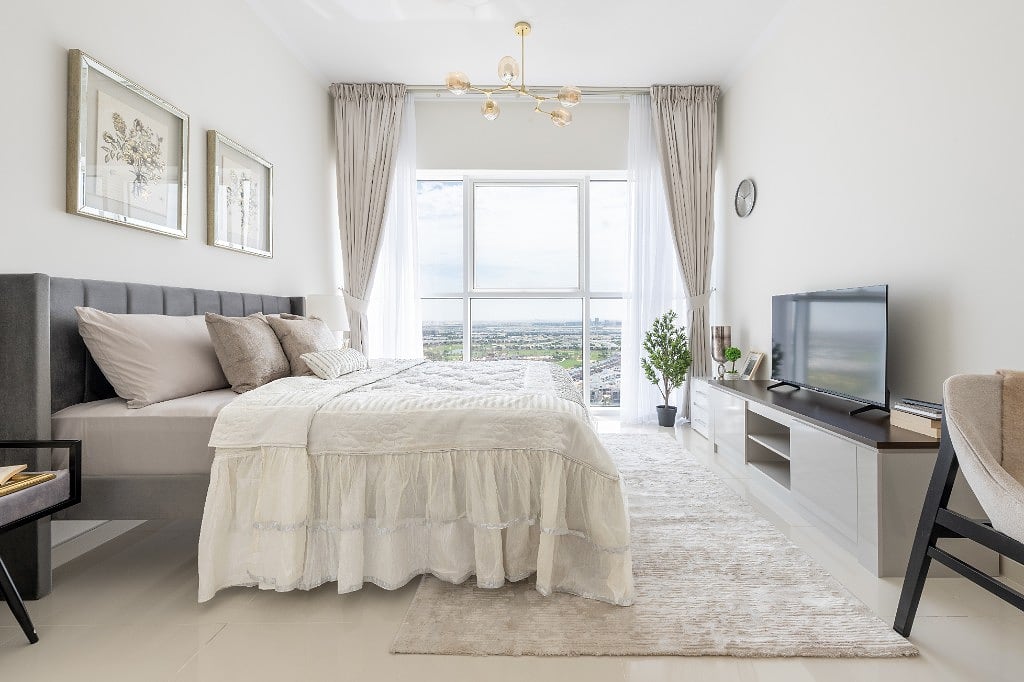 A golf-view apartment in Damac Hills, a grey upholstered bed against a floor-to-ceiling window onto the golf course, with sheer linen and taupe drapery, a gold glass chandelier and a white media console