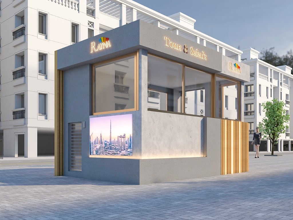 A side view of the JBR kiosk, a glazed concrete pavilion with illuminated fascia signage and a backlit Dubai-skyline panel set into the lower wall, on a paved plaza