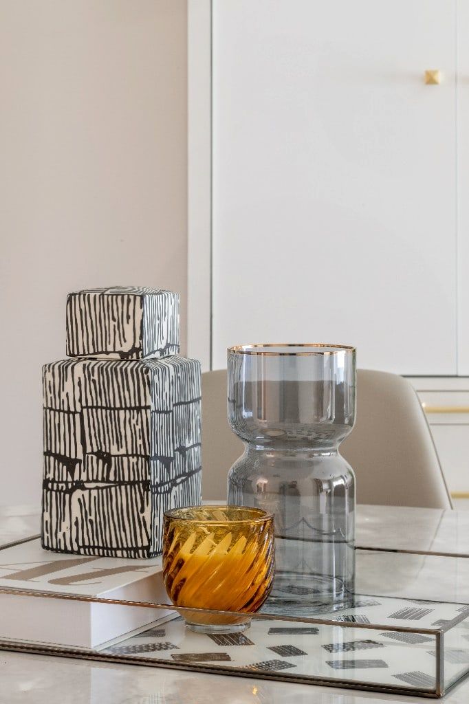Styling vignette on the living room coffee table, a striped black-and-cream lidded jar, a tall smoky-glass vase with a brass rim and a fluted amber tealight on a mirrored tray