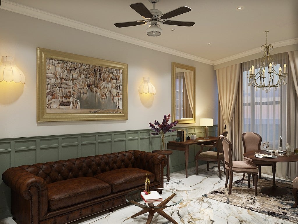 A suite living area with a deep brown button-tufted leather chesterfield against sage-green wainscoting, a gold-framed artwork, a writing desk and a round dining table under a crystal chandelier