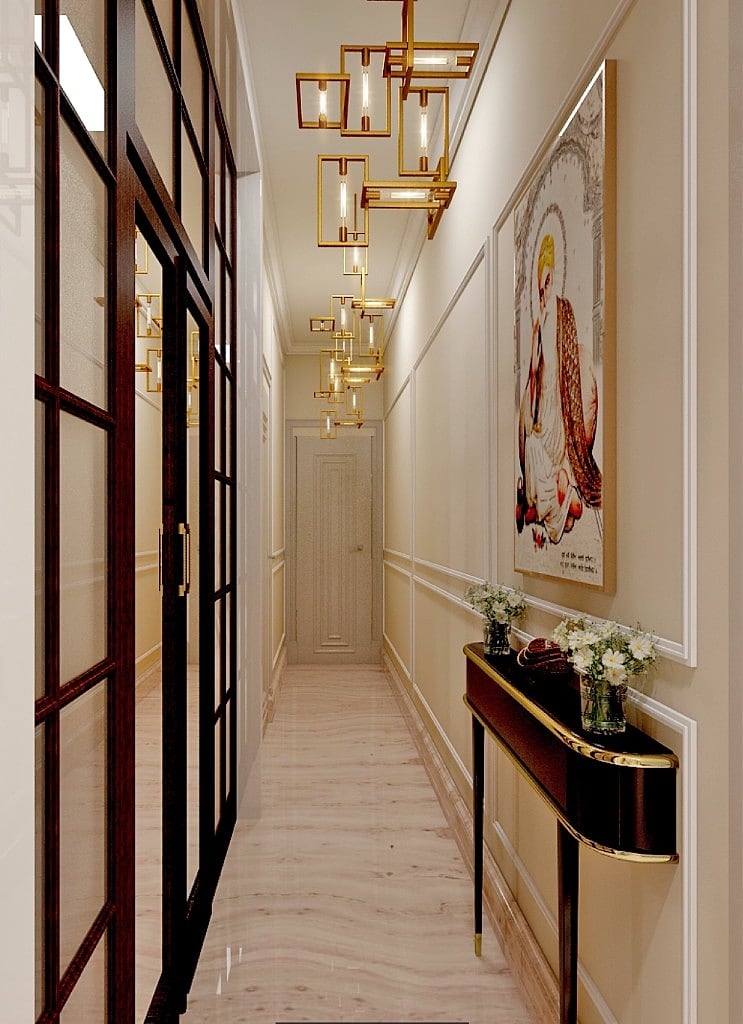 A gallery-like corridor lit by a cascade of brass-framed pendant lights, with panelled walls, a black-framed glass partition, a curved console with white flowers and a piece of Indian art